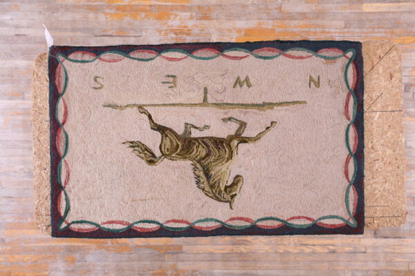 Hooked beige rug with stylized upside-down brown horse motif and colored compass letters on wood floor.