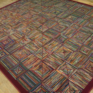 A vibrant geometric rug made of colorful woven strips, framed by a burgundy border on light wood planks.