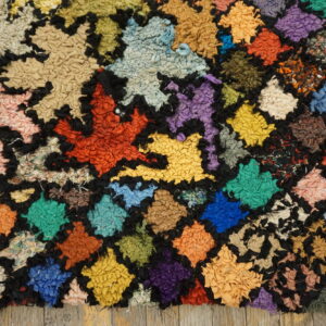 A multicolored, chunky-textured rag rug with a geometric grid pattern rests on worn wood flooring.