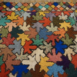 Colorful textured rug featuring large leaf shapes and a checkered border on rustic wooden floorboards.
