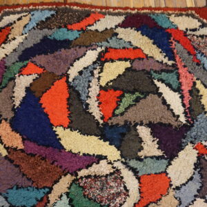 A shaggy, multicolored hooked rug with an abstract patchwork design rests on distressed wood flooring.