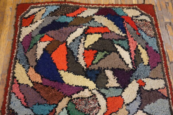A shaggy, multicolored hooked rug with an abstract patchwork design rests on distressed wood flooring.