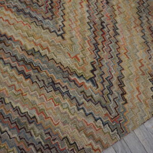 Textured rug featuring dense multicolored zigzag patterns in muted earth tones on grey wooden flooring.