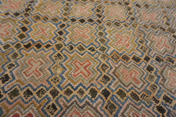 Textured rug surface featuring geometric zigzag and cross motifs in muted blue, pink, gold, and dark green.