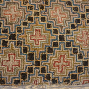 Multicolored textured rug with geometric crosses and dark squares resting on light weathered wood flooring.