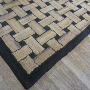 Textured rug corner featuring black and multi-tonal beige interlocking rectangles over distressed plank flooring.