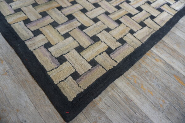 Textured rug corner featuring black and multi-tonal beige interlocking rectangles over distressed plank flooring.