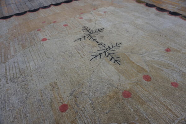 Muted beige textured rug with a black leaf motif and coral spots, lying on gray wooden floorboards.