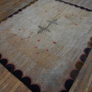Textured beige and grey rug with dark central leaves, pink dots, and a scalloped border on worn wood floors.