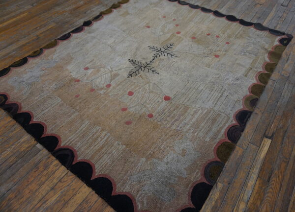 Textured beige and grey rug with dark central leaves, pink dots, and a scalloped border on worn wood floors.