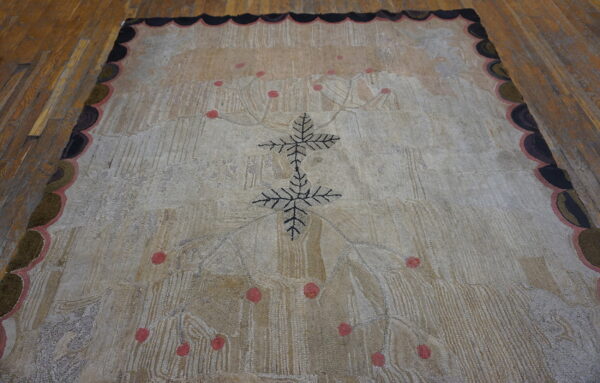 Textured beige rug with a black leafy center, pink dots, and a multicolored scalloped border on wood flooring.