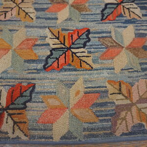 A textured hooked rug displays multicolored leaf patterns in orange, tan, and green on a striated blue background.