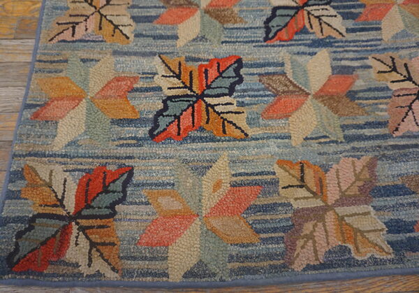 A textured hooked rug displays multicolored leaf patterns in orange, tan, and green on a striated blue background.