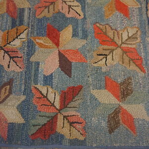Blue hooked rug featuring large textured autumn leaves and geometric stars in coral, tan, and brown tones.