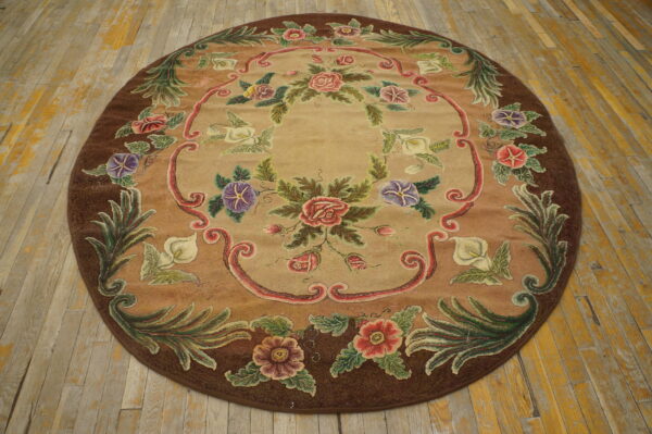 Round floral rug in tan, brown, pink, and green colors displayed on worn light hardwood plank flooring.