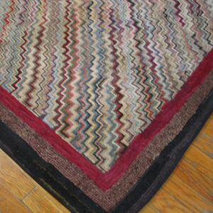 Multicolored zigzag rug corner with layered red, brown, and black borders rests on light wood planks.