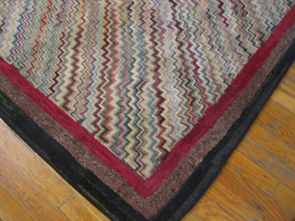 Multicolored zigzag rug corner with layered red, brown, and black borders rests on light wood planks.