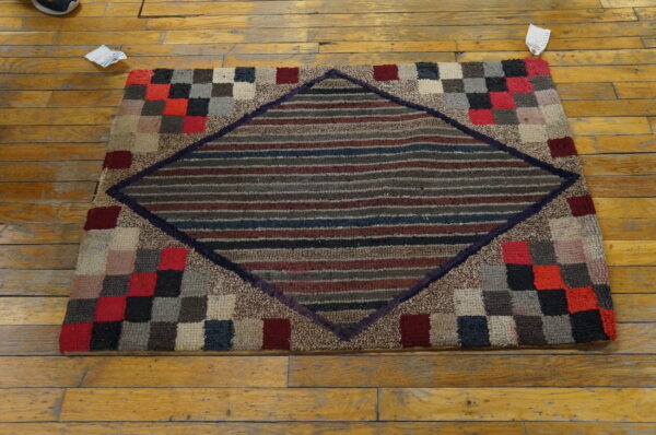 Textured multicolored rug displaying a striped central diamond framed by a checkered block border on wood flooring.