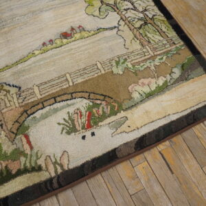 Hooked rug with a pastoral scene of a bridge and trees in muted greens and browns on rustic wood flooring.