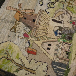Beige, green, and brown looped rug depicting a stylized windmill and village scene on wood floor.