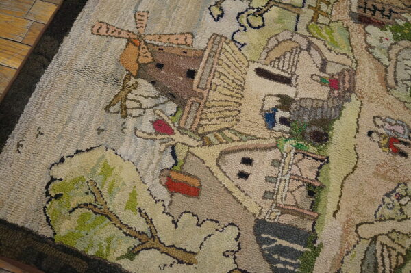 Beige, green, and brown looped rug depicting a stylized windmill and village scene on wood floor.