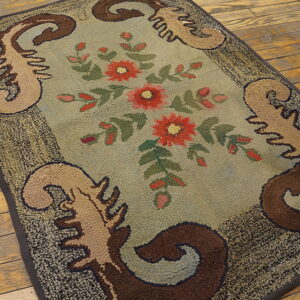 Hooked rug with red florals and brown scrolls on a sage field, placed on distressed wood plank flooring.