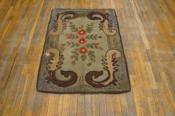 Hooked rug with red flowers and brown scrolls on a blue-green center, displayed on distressed wood floors.