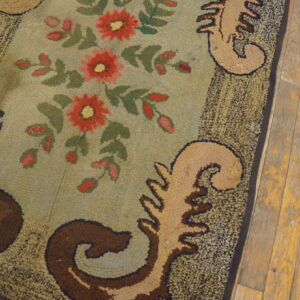 Textured rug with a sage green field, red florals, and tan scrollwork bordered by mottled dark brown on wood flooring.
