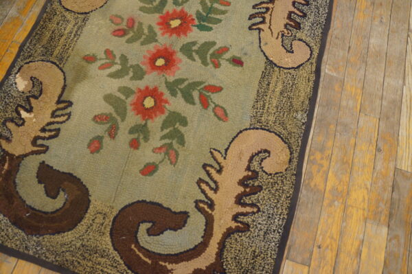 Textured rug with a sage green field, red florals, and tan scrollwork bordered by mottled dark brown on wood flooring.