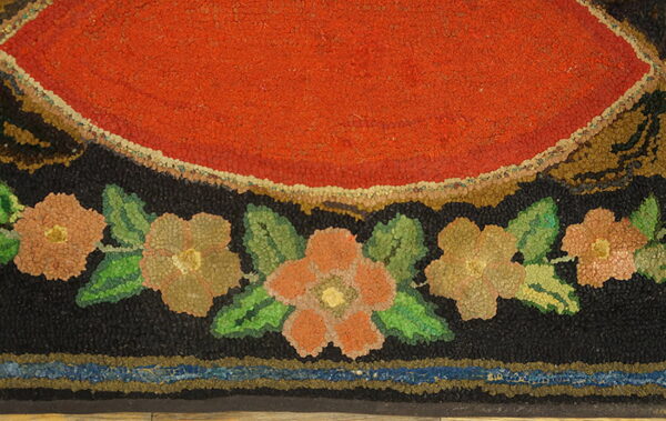 Hooked rug detail featuring a bright red center, black field, and a border of green leaves and multicolored flowers.