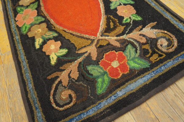 Hooked rug with a red center, multicolored floral patterns, black background, and striped border resting on wood planks.