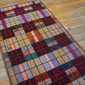 Multicolored checkered rug with varying textures in squares of maroon, purple, and tan on weathered wood flooring.