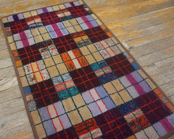 Multicolored checkered rug with varying textures in squares of maroon, purple, and tan on weathered wood flooring.