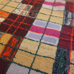 Multi-toned, looped pile rug featuring a vivid patchwork grid in shades of red, pink, beige, and blue.