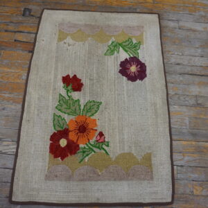 Beige loop pile rug featuring large red, orange, and purple flowers rests on a worn wooden floor.