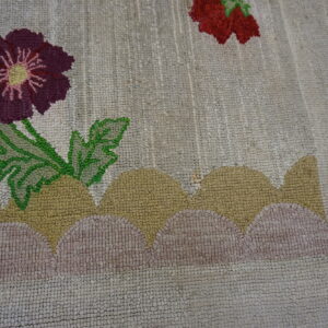 A looped rug with a beige background displays purple, red, and green floral designs on stylized tan and pink hills.