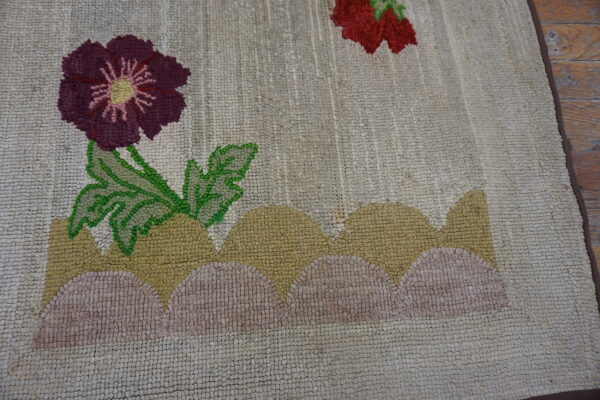 A looped rug with a beige background displays purple, red, and green floral designs on stylized tan and pink hills.