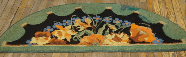 Hooked semicircular rug with orange and blue flowers on black, surrounded by a scalloped green border on wood planks.