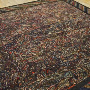 Multicolored hooked rug showing a dense, dark abstract pattern and striped border on light wood flooring.