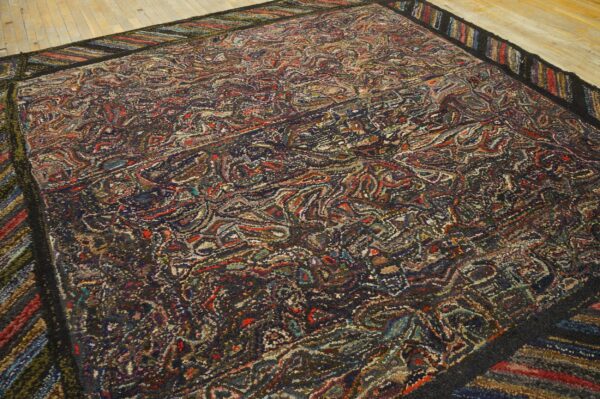 Multicolored hooked rug showing a dense, dark abstract pattern and striped border on light wood flooring.