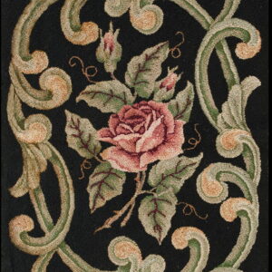 Close up of a textured rug showing a pink rose and green scrolling vines on a deep black background.