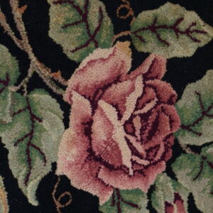 Textured close-up of a floral rug featuring a pink rose and green leaves on a black ground.