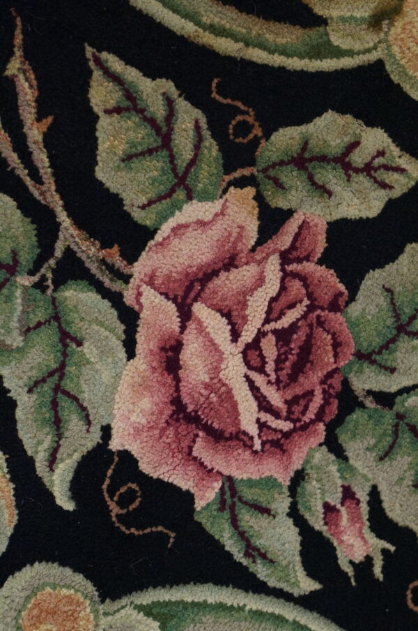 Textured close-up of a floral rug featuring a pink rose and green leaves on a black ground.