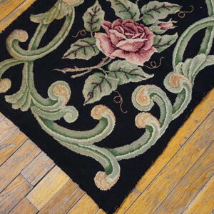 Textured black rug featuring a large pink rose and thick green scrolls on worn hardwood flooring.