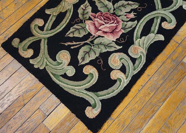 Textured black rug featuring a large pink rose and thick green scrolls on worn hardwood flooring.