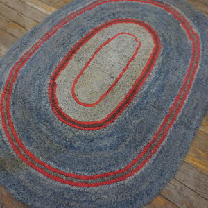 Oval blue and gray hooked rug with concentric red stripes on distressed wood flooring.