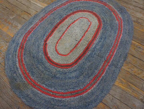 Oval blue and gray hooked rug with concentric red stripes on distressed wood flooring.