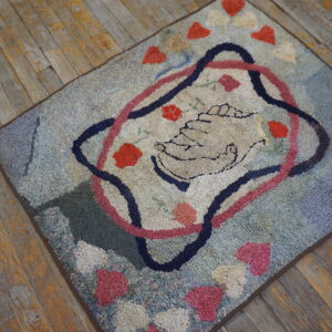 Nubby blue and pink hooked rug with a central clasped hands motif on worn wood planks.