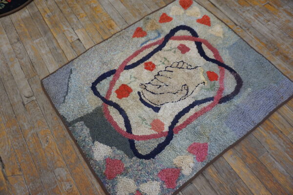 Nubby blue and pink hooked rug with a central clasped hands motif on worn wood planks.