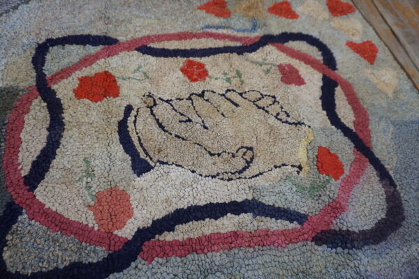 Textured, multi-colored rug featuring a central motif of two hands and scattered red floral accents on a wood floor.